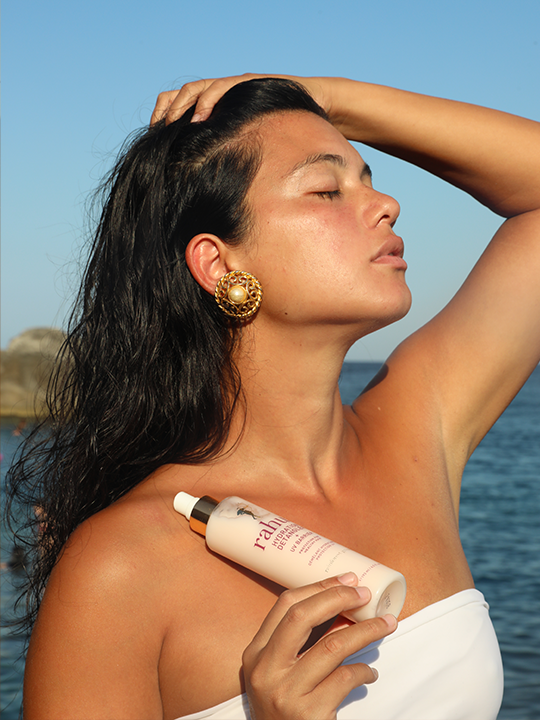 model posing in the sun by the seaside with a bottle of rahua hydration detangler and uv barrier|variant:65-oz