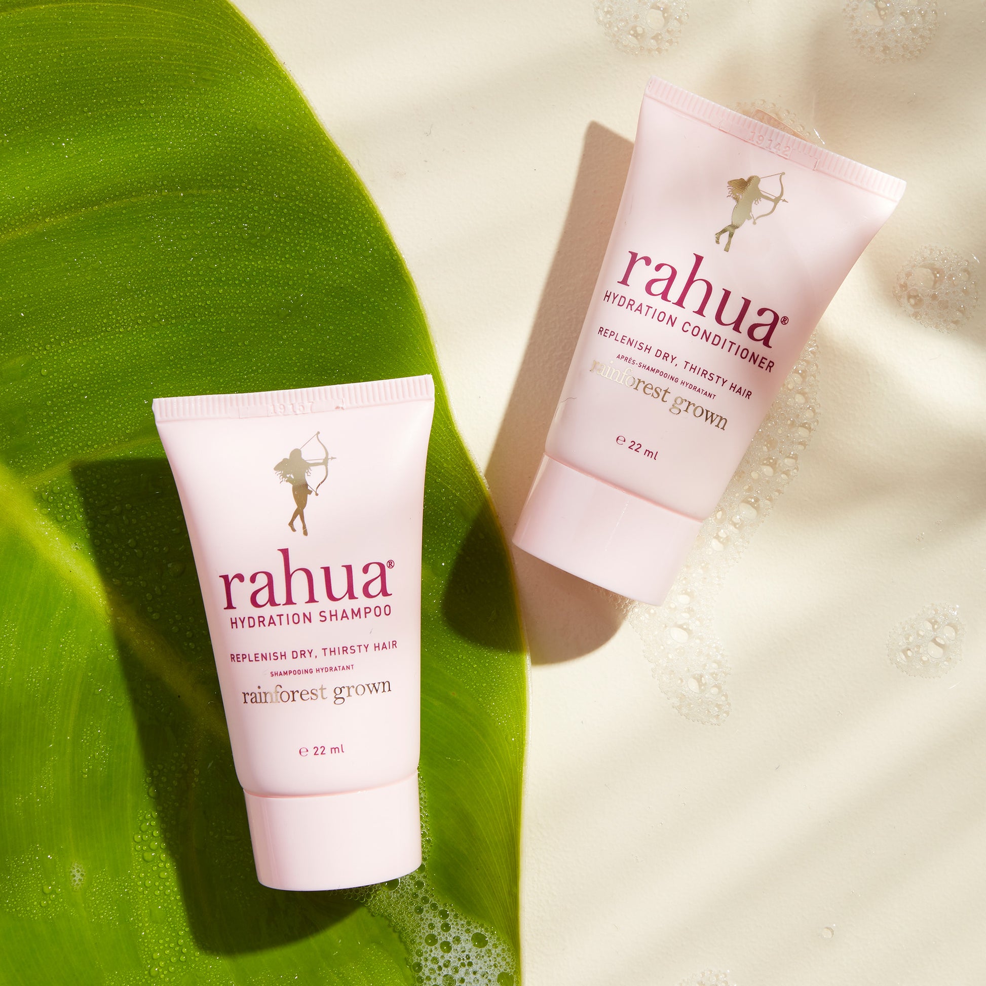 two tubes of deluxe mini size rahua hydration conditioner and shampoo on a soapy beige background with a leaf on the left side|variant:074-oz